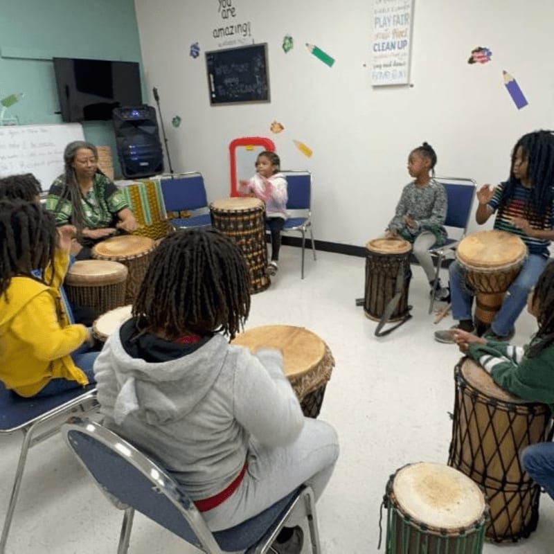 Children and an adult are gathered in a circle, playing drums in a room with colorful decorations on the walls.