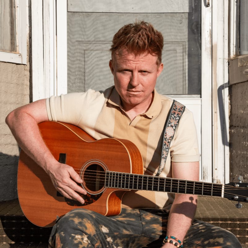 A man with red hair sits on a porch with an acoustic guitar, while a screen door is visible behind him.