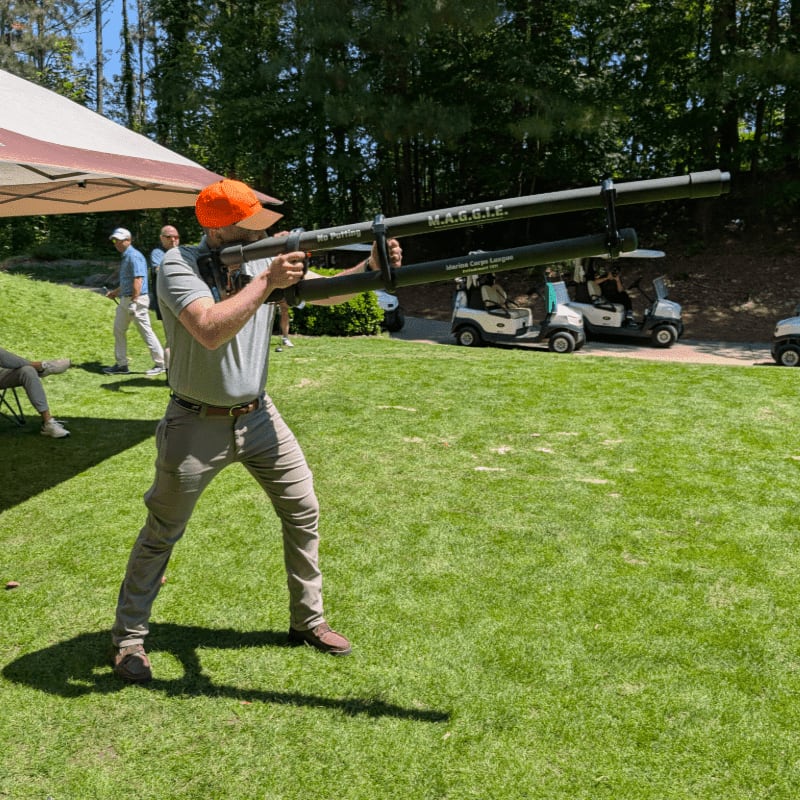 A man in an orange hat aims a large contraption on a grassy field with golf carts and trees in the background.
