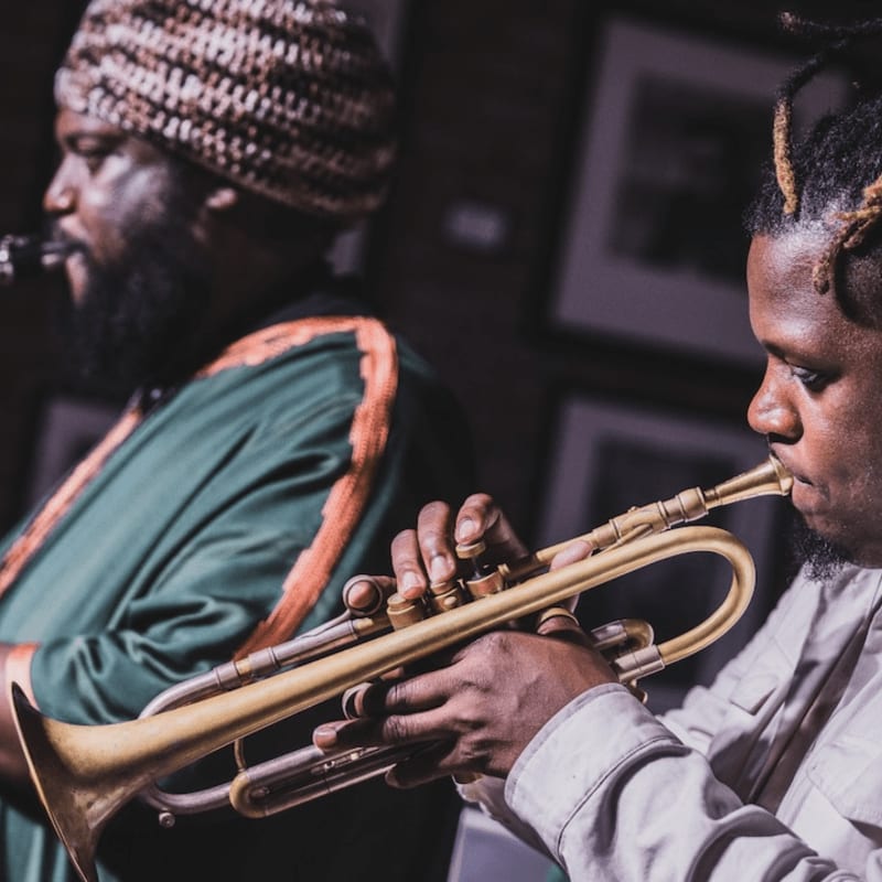 A man in a light-colored shirt plays a trumpet in the foreground, while another man in a green and orange robe plays a clarinet in the blurred background.