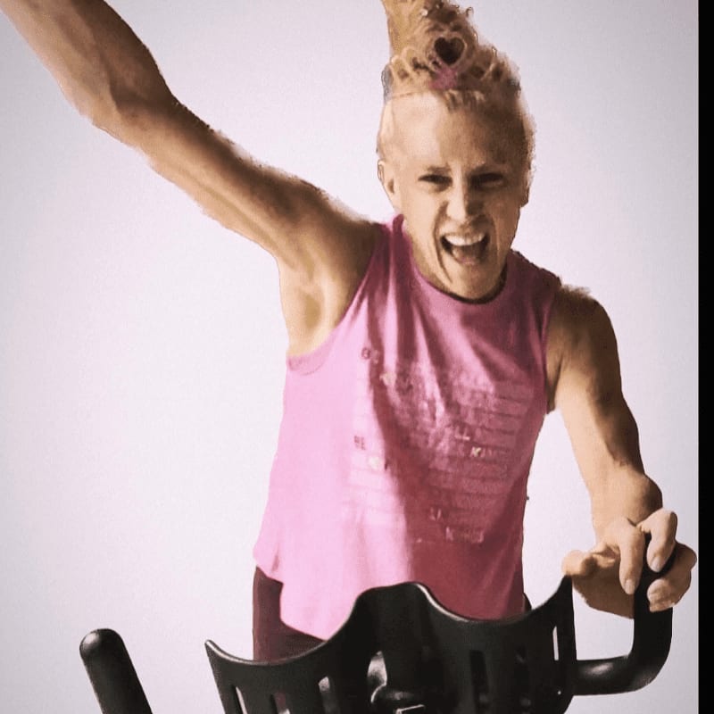 A woman with a spiky hairstyle and a pink tank top exuberantly rides a stationary bike against a plain white background.