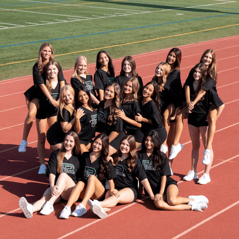 A group of cheerleaders in black uniforms pose on a red track with a blurred green football field in the background.