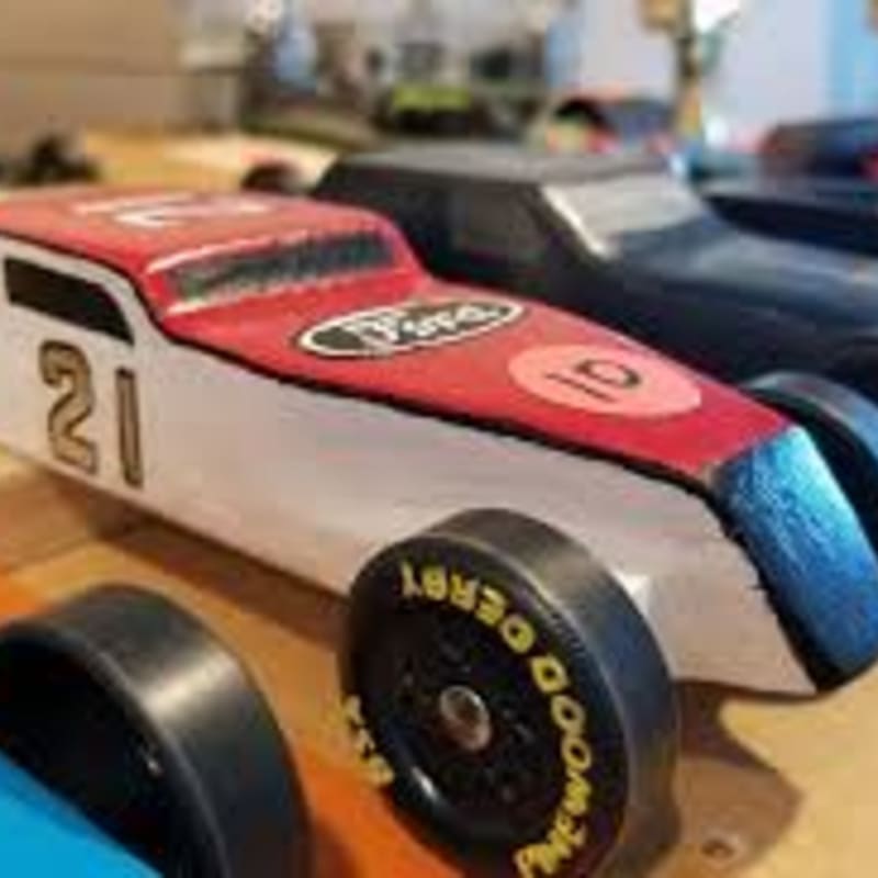 A red and white pinewood derby car with the number 21 is in the foreground, with other cars and a blurred background.