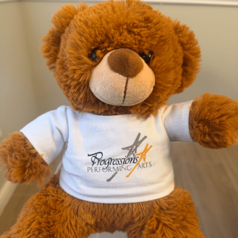 A fluffy brown teddy bear wearing a white t-shirt with a logo stands against a blurred background of a wall and floor.