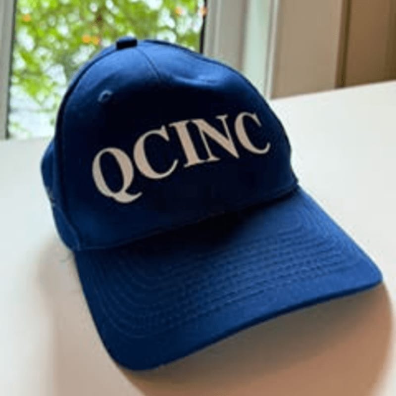 A blue baseball cap with "QCINC" written in white is in the foreground, with a blurred background of a window and greenery.