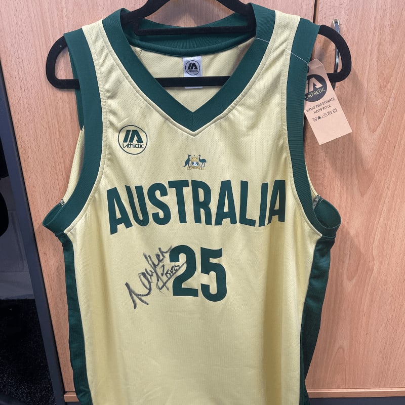 A yellow and green Australia basketball jersey with the number 25 and an autograph hangs on a hanger in front of a wooden cabinet.