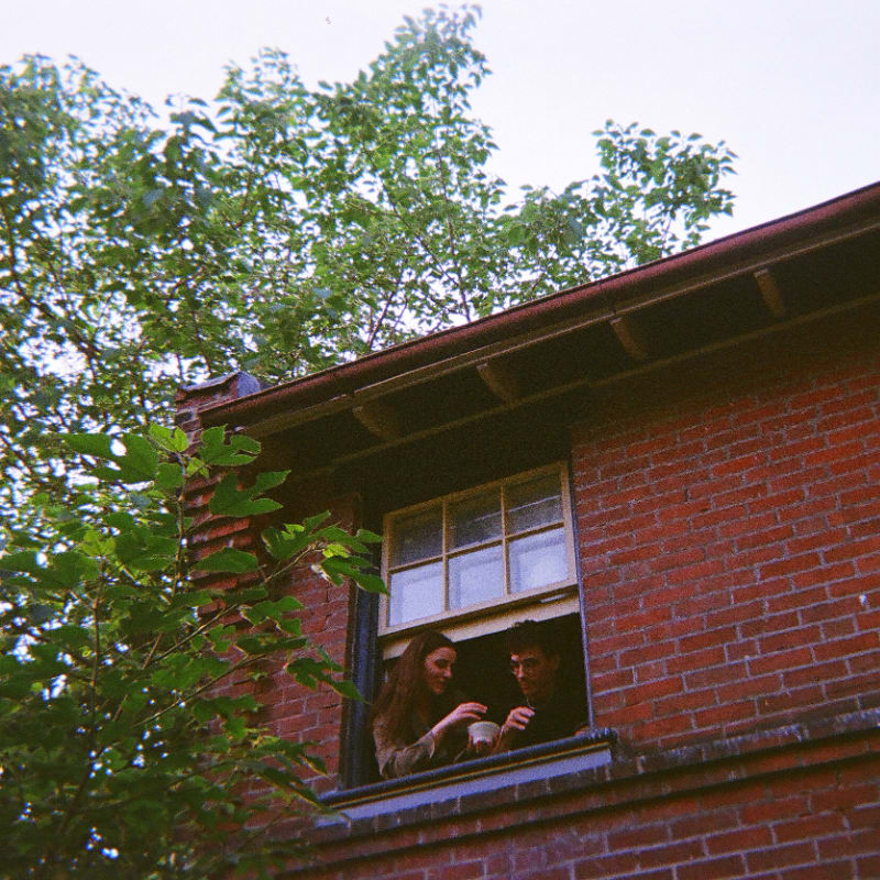 A young couple shares a moment by a window framed by lush green trees and a red brick building.