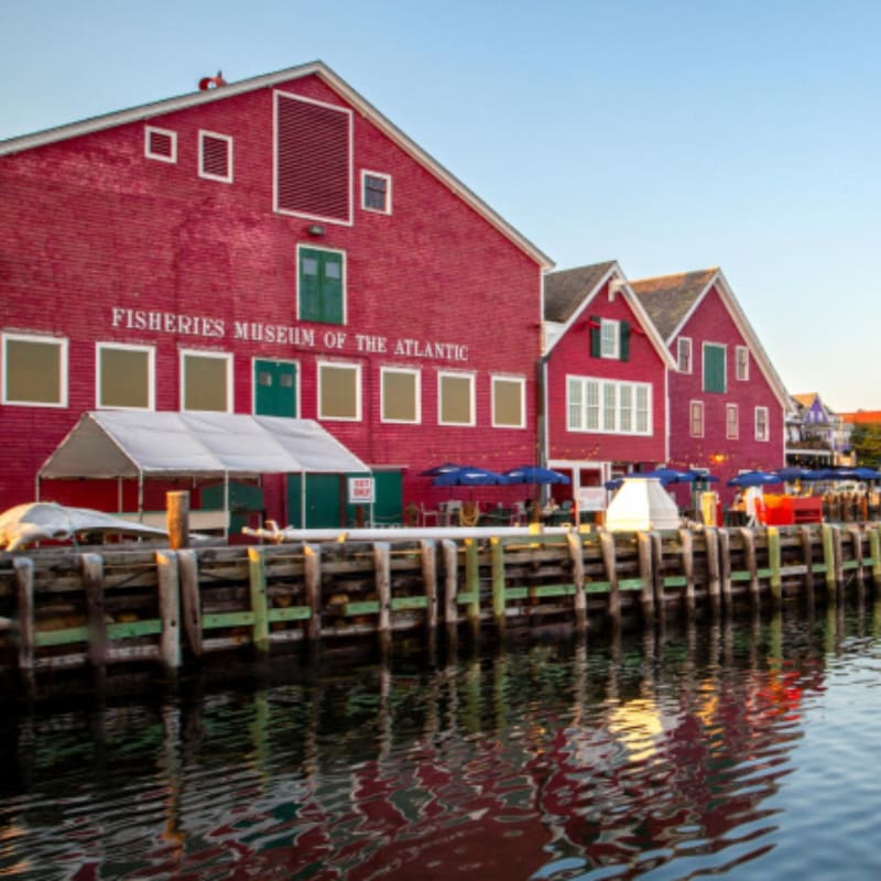A bright red building labeled "Fisheries Museum of the Atlantic" stands on a wooden pier overlooking calm water reflecting the sky and buildings.