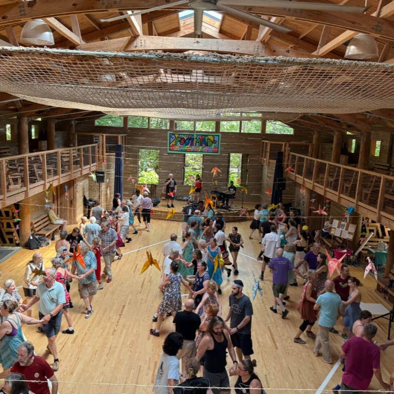 People dance in a large wooden hall with a band playing on a stage in the background.