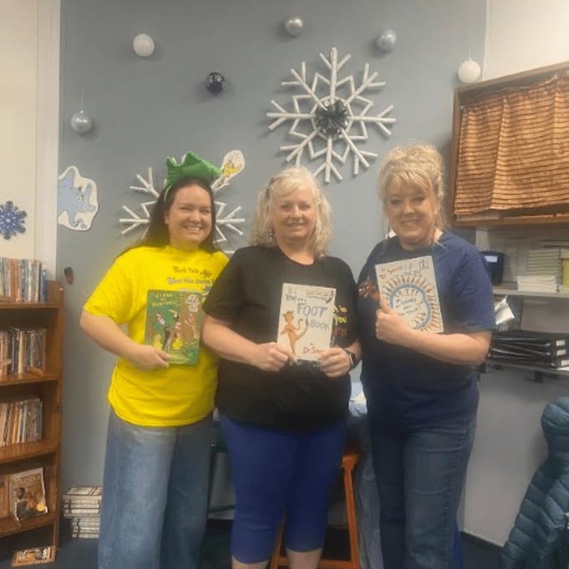 Three women stand in front of a wall decorated with snowflakes and Dr. Seuss characters, holding books.