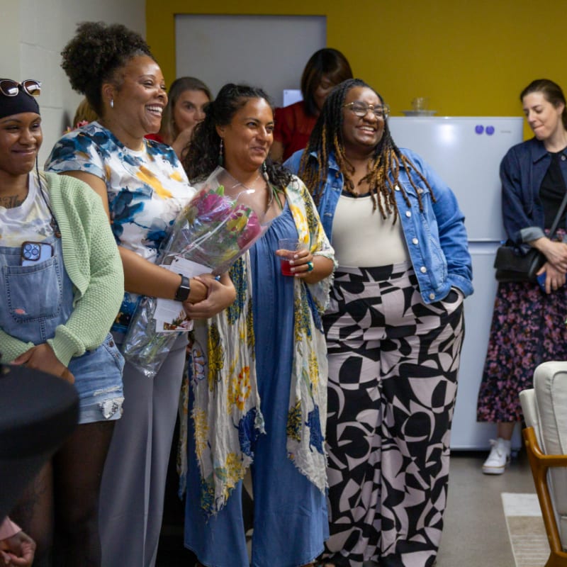 A group of women smile and laugh in the foreground, holding a bouquet of flowers, while a yellow wall and a refrigerator are visible in the background.