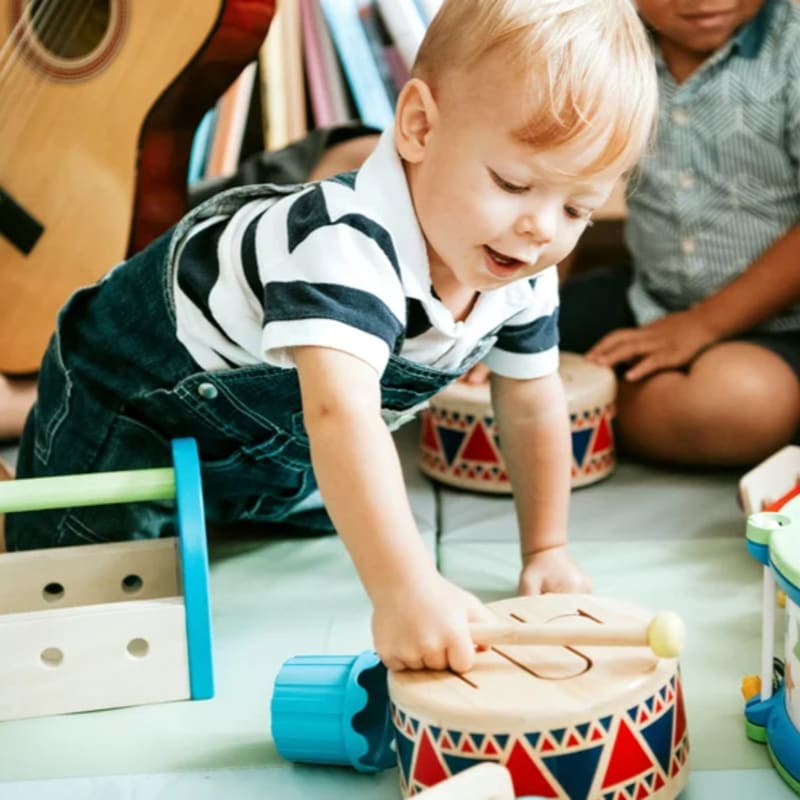 A young boy in a striped shirt and denim overalls plays with a toy drum in the foreground, while other children and musical instruments are visible in the blurred background.