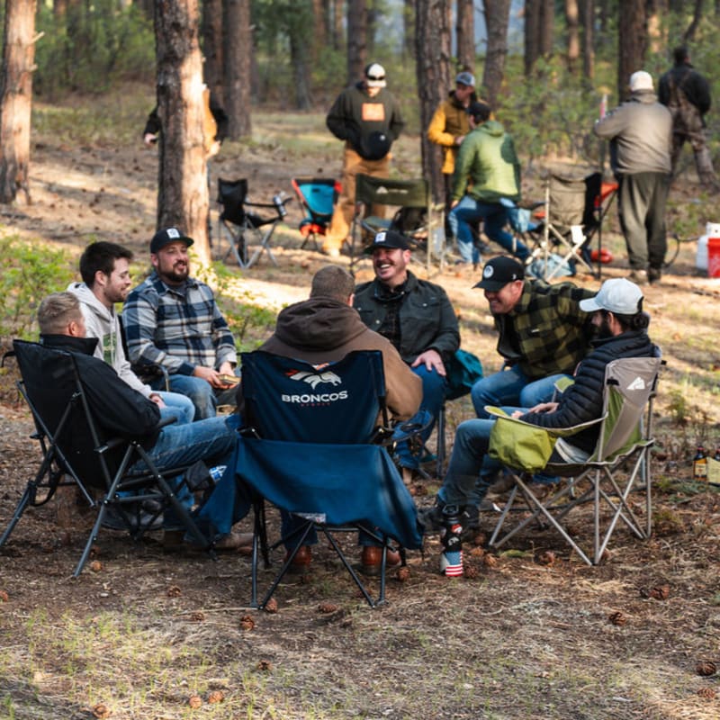 A group of men in casual attire sit in camping chairs in a forest clearing, with more people and camping gear visible in the background among the trees.