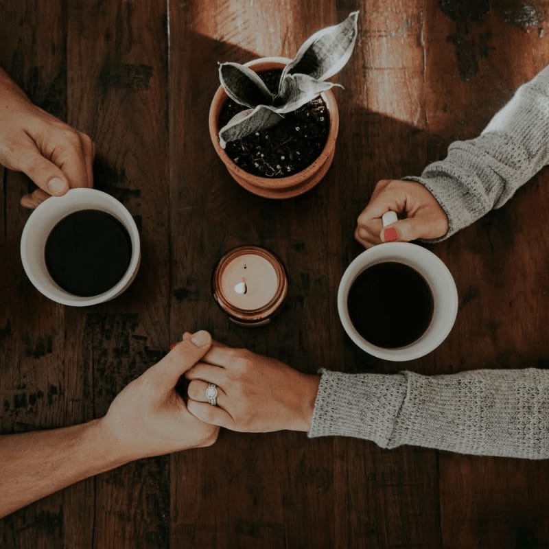 Two people hold hands over a wooden table, with cups of coffee and a small potted plant in the background.