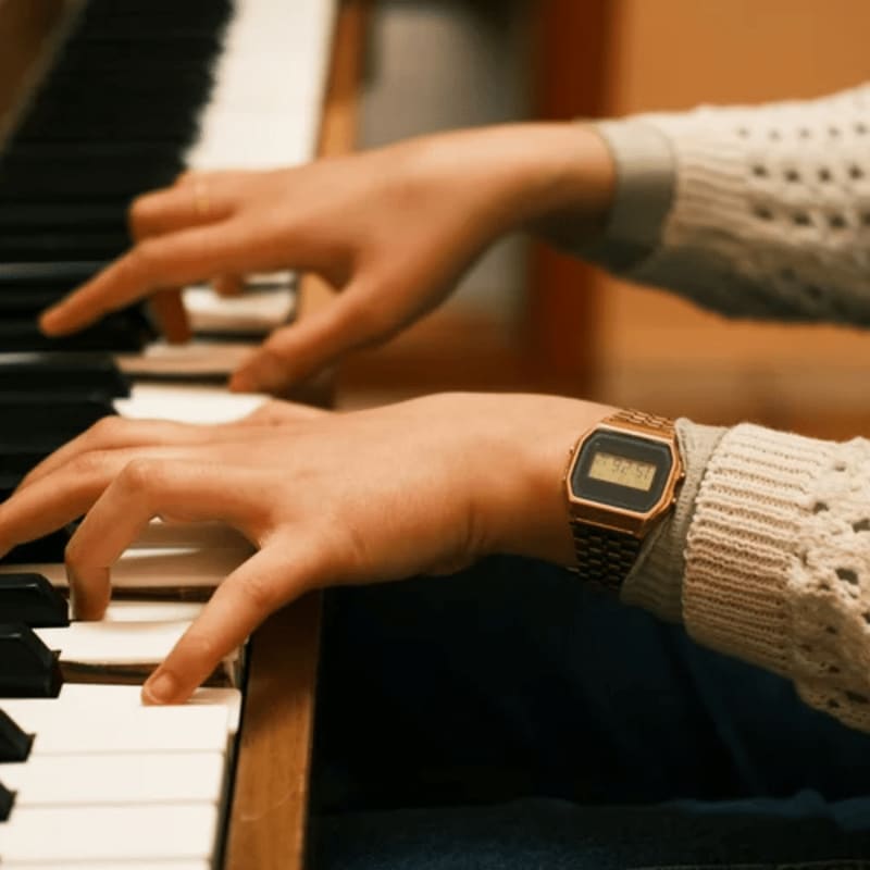Hands with a watch play piano keys in the foreground, with a blurred warm-toned background.