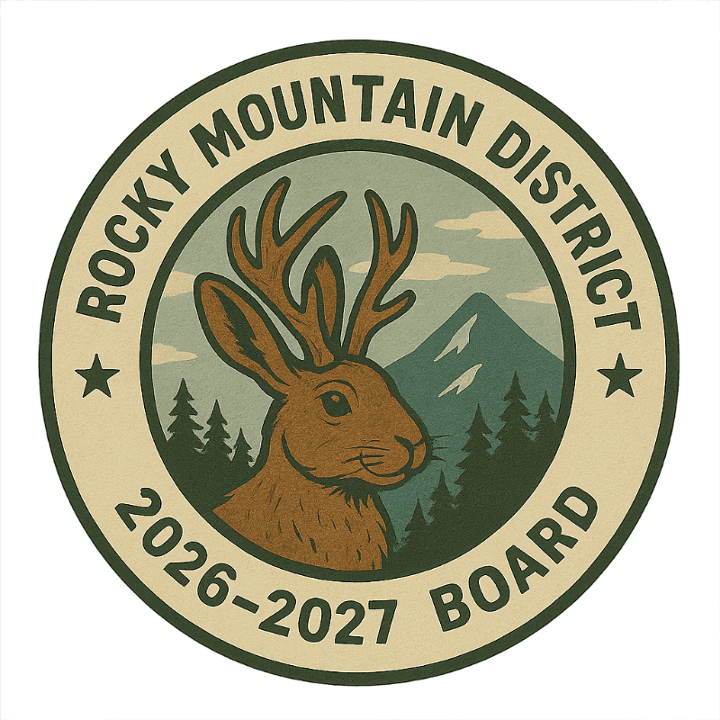 A jackalope with antlers is featured in the foreground, set against a background of pine trees and a snow-capped mountain under a cloudy sky, all within a circular emblem that reads "ROCKY MOUNTAIN DISTRICT 2026-2027 BOARD".