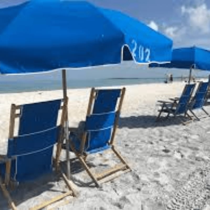 Blue beach chairs and umbrellas sit on a sandy shore with the ocean and a hazy sky in the background.