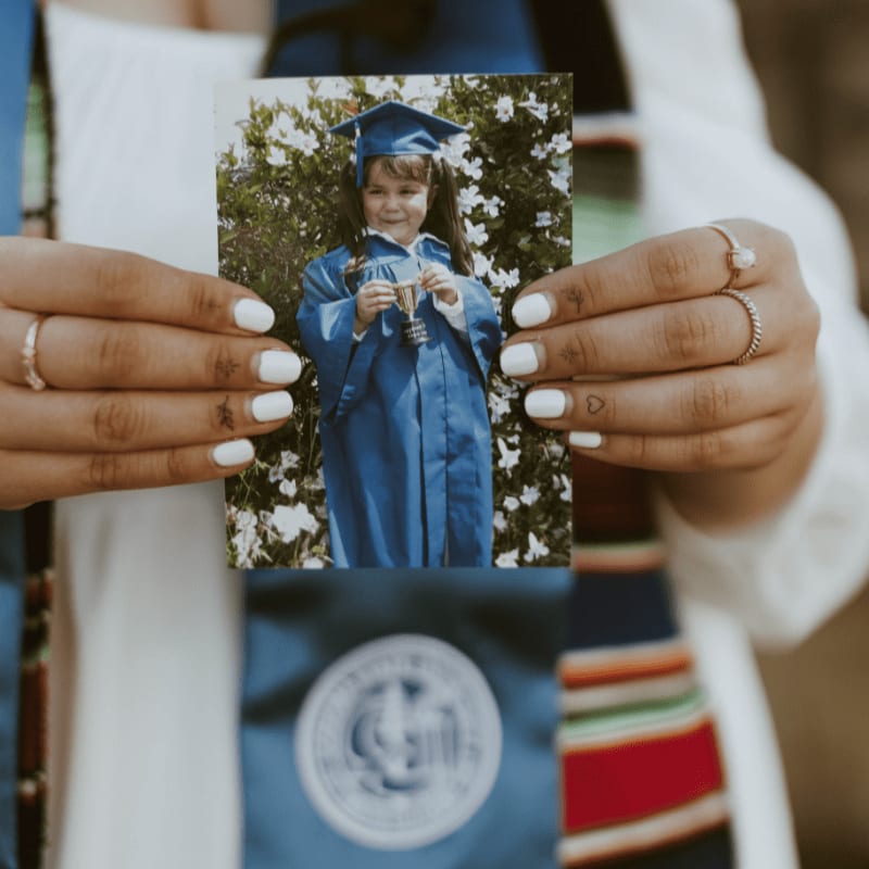 Hands with white nail polish hold a photograph of a young girl in a blue graduation gown, with a blurred background of white flowers and a graduation stole visible in the foreground.