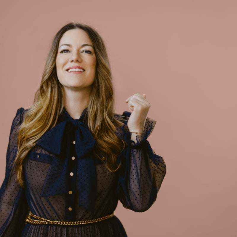A woman with long, wavy brown hair smiles and raises her fist in front of a plain pink background.