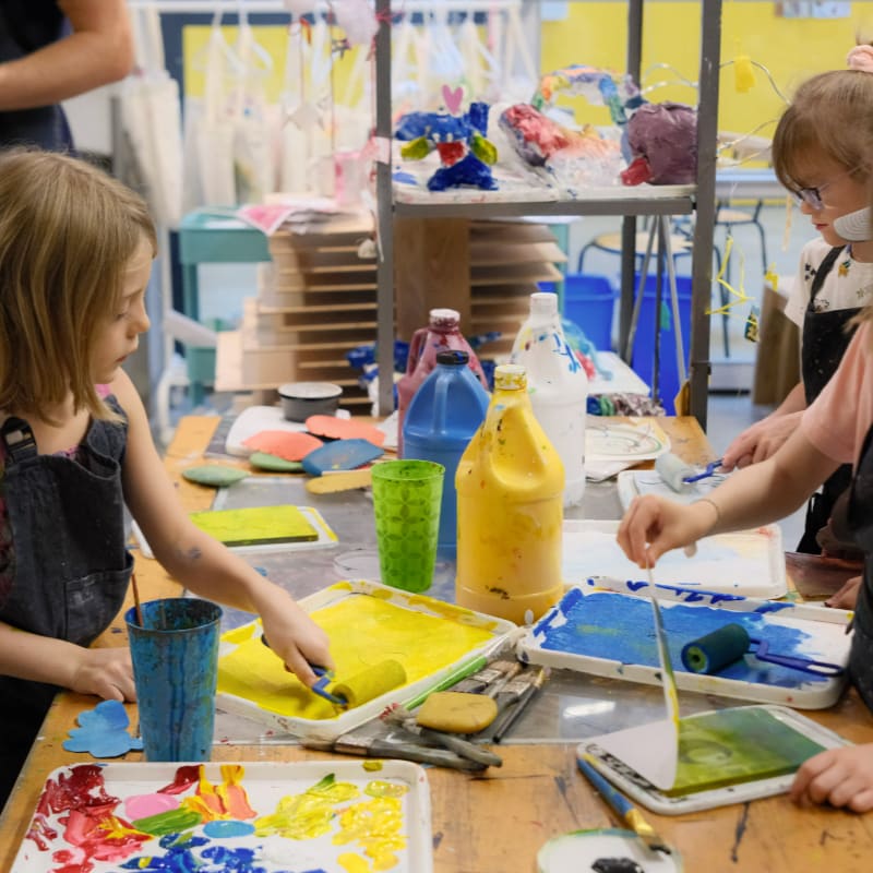 Children in the foreground are engaged in a colorful art project with paint rollers and trays, while the background reveals shelves filled with various art supplies and creations.
