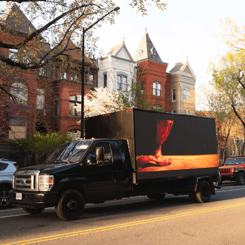 A black truck with a large LED screen displaying a close-up of a foot is parked on a street in front of ornate, multi-story houses.