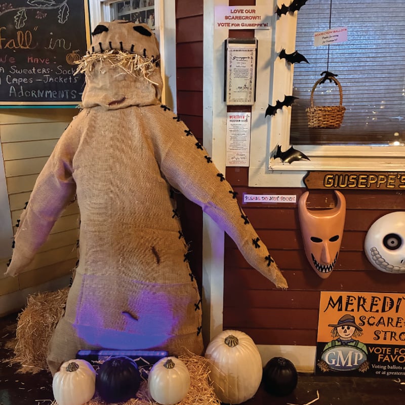 A burlap scarecrow with straw hair stands in the foreground, surrounded by pumpkins, while a decorated window with Halloween cutouts and signs is visible in the background.