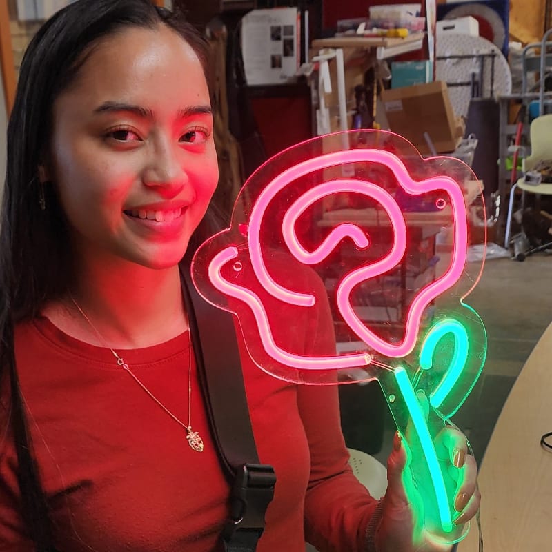 A smiling young woman in the foreground holds a neon rose sign with pink petals and a green stem, while the background shows a cluttered workshop.