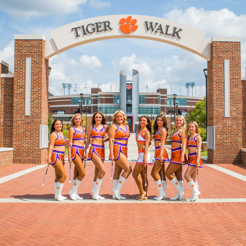 Eight young women in orange and white cheerleading outfits stand with batons on a brick pathway in front of a large stadium under a "Tiger Walk" archway.
