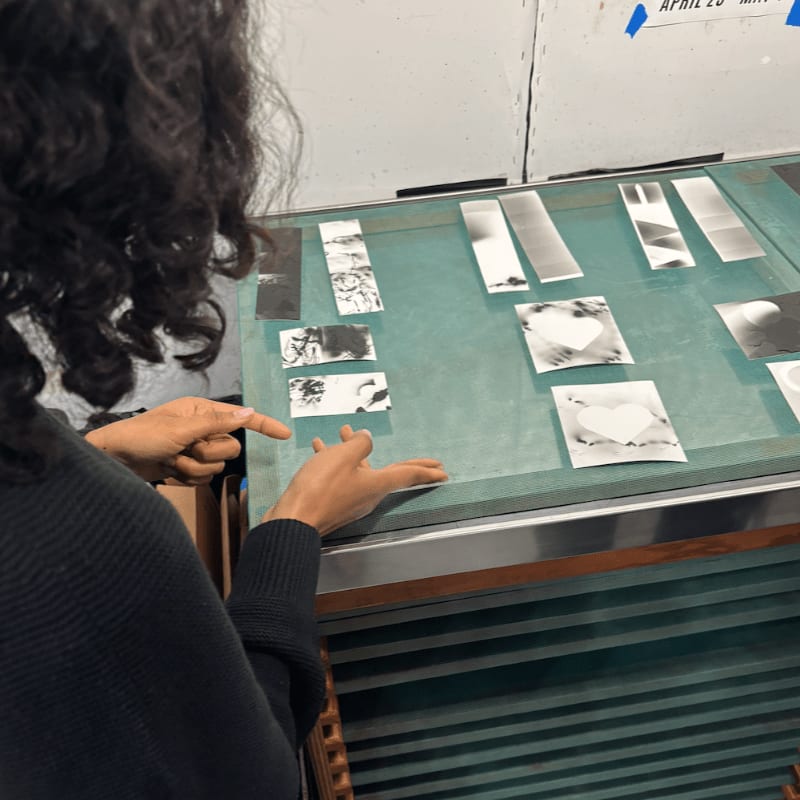 A person's hands point to various black and white photographs laid out on a teal surface, with a white wall and a blue tape marking visible in the background.