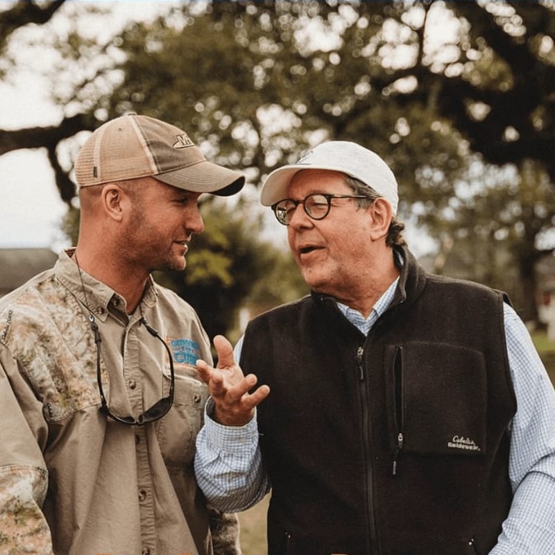 Two men in casual outdoor attire converse in the foreground, with a blurred background of trees and foliage.