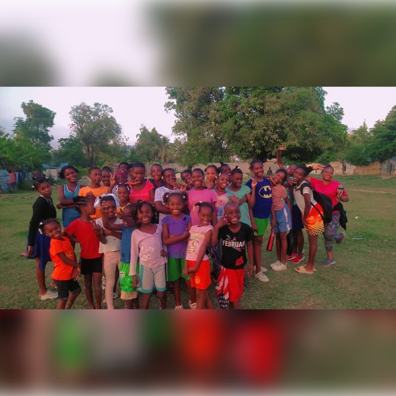 A large group of smiling children, dressed in colorful clothing, pose for a photo in a grassy field with trees and buildings in the background.