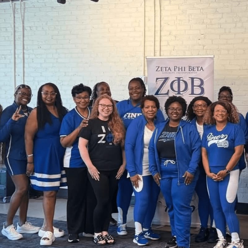 A group of women in blue and white Zeta Phi Beta attire pose in front of a banner, with a brick wall visible in the background.
