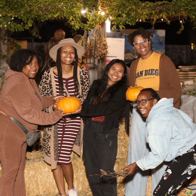 Five smiling women pose with pumpkins in the foreground, while a dimly lit, rustic background with hay bales and string lights suggests an outdoor autumn event.