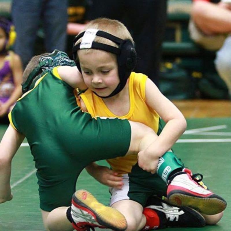 Two young boys in wrestling gear grapple on a green mat, with blurred spectators in the background.