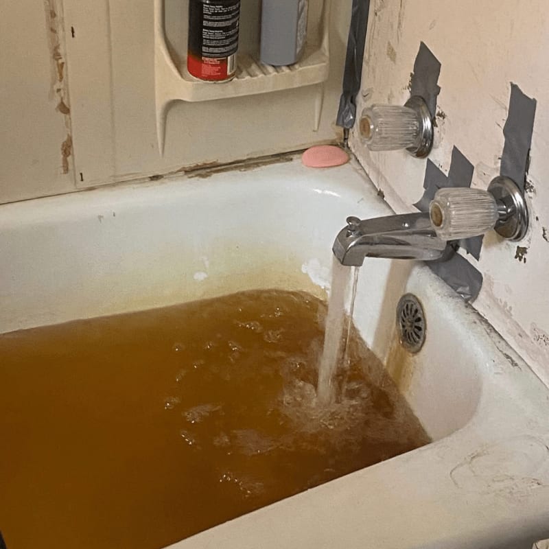A bathtub is filling with murky brown water, with a soap bar and bottles visible on the shelf above, and peeling paint and taped-up fixtures on the wall behind.