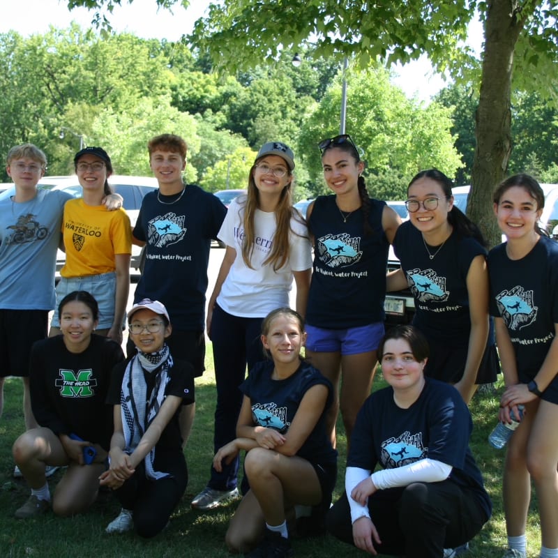 A group of young people pose for a photo outdoors with trees and a vehicle in the background.