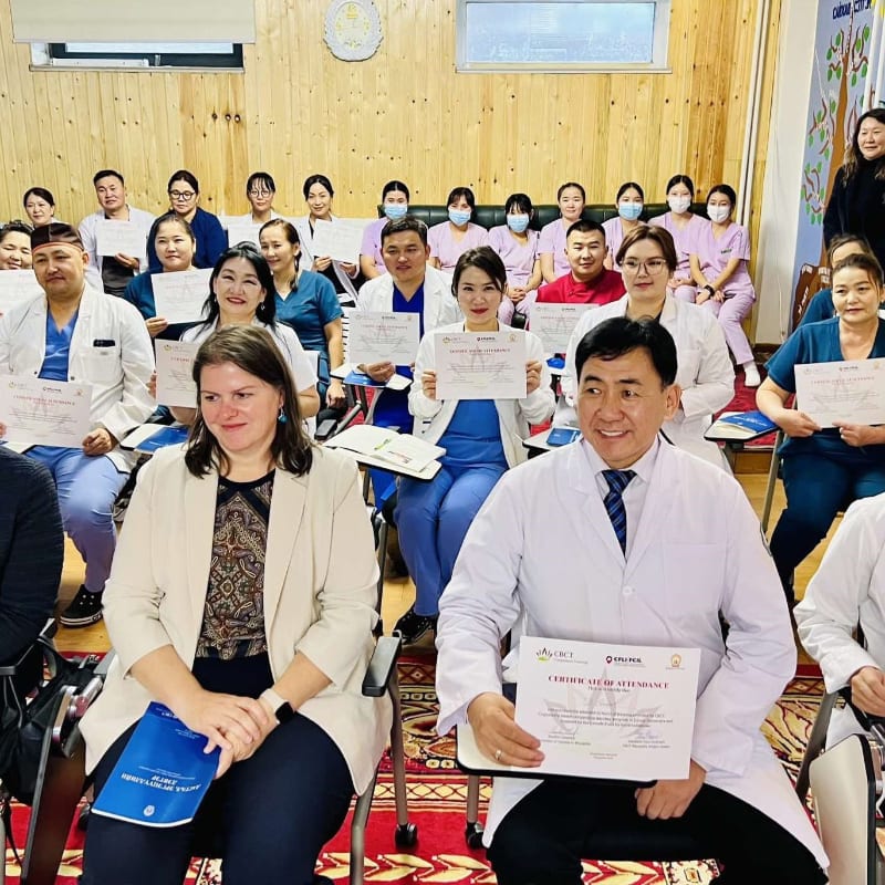 Two women in the foreground are seated with certificates, while a group of people in medical attire are seated behind them in a room with wooden walls.