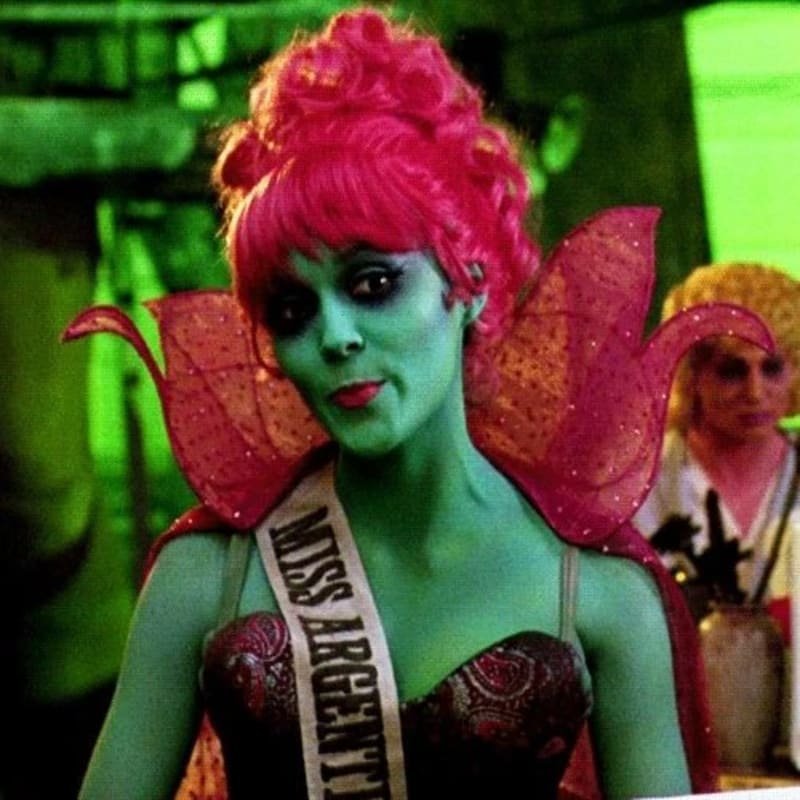A woman with green skin, bright pink hair, and a "Miss Argentina" sash stands in the foreground, while a blurred figure with yellow hair is visible in the green-lit background.