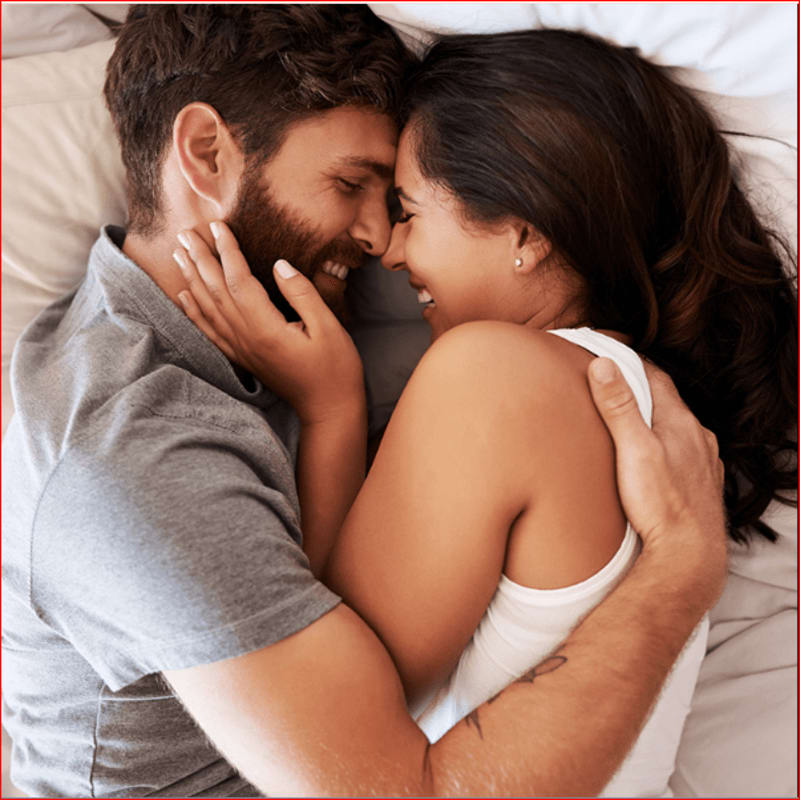 A couple embraces tenderly on a white bed, their faces close and smiling.