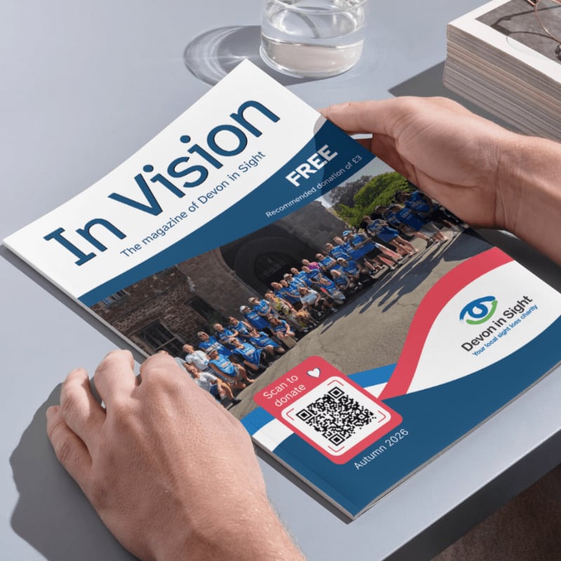 Hands hold an open magazine titled "In Vision" with a QR code for donations in the foreground, while a stack of papers and a glass of water rest in the background.