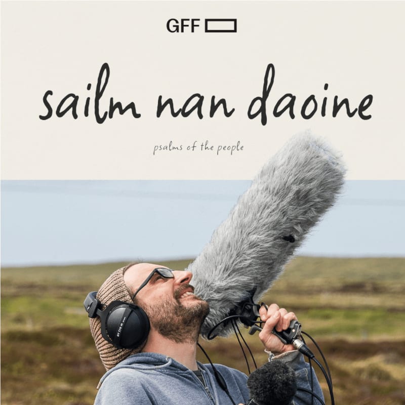 A man wearing headphones and a beanie holds a furry microphone in the foreground, with a blurred landscape and sky in the background.