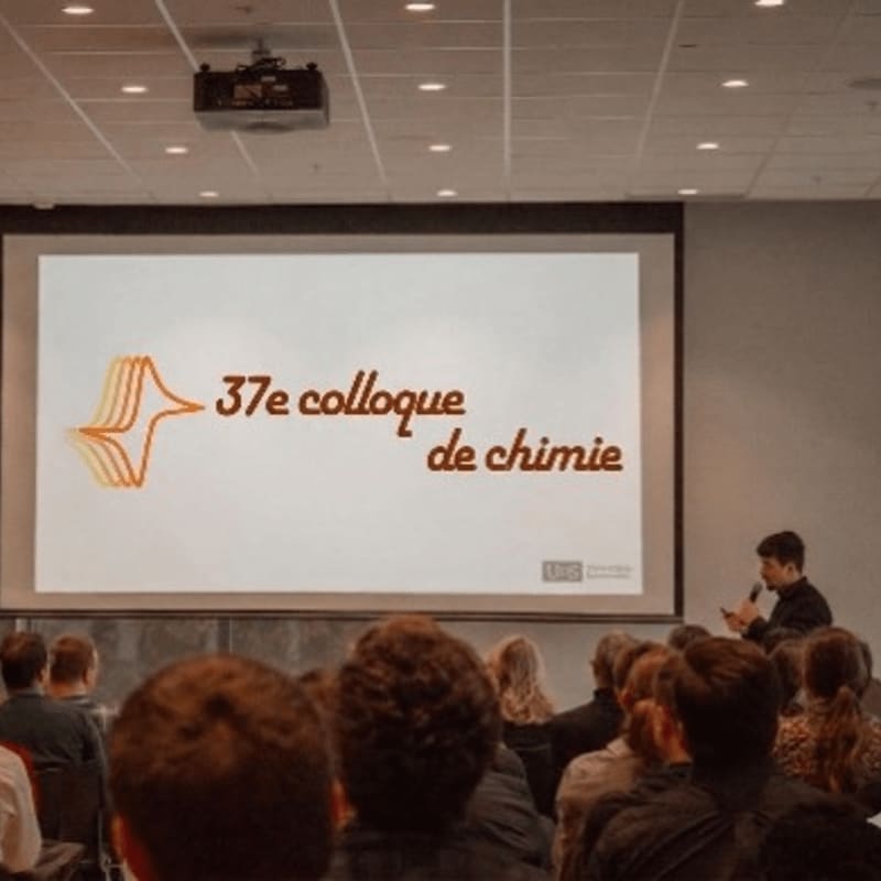 A speaker presents to an audience in a conference room, with a screen displaying "37e colloque de chimie" in the background.