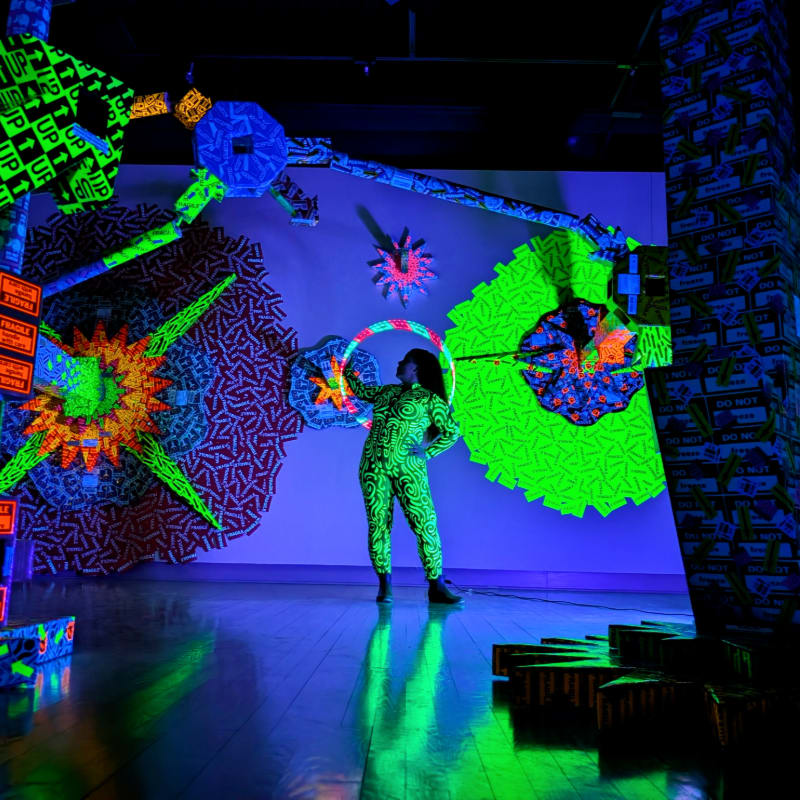 A person in a neon green patterned suit stands in a room filled with glowing, geometric art installations and projections.
