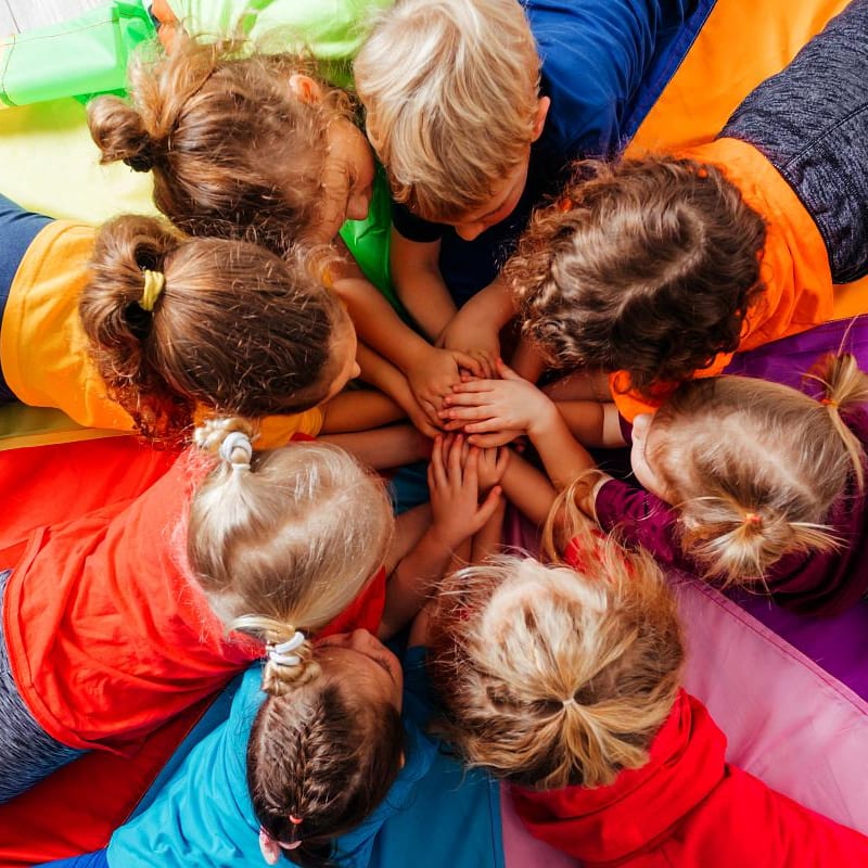A group of children with their hands clasped in the center are lying on a colorful parachute.