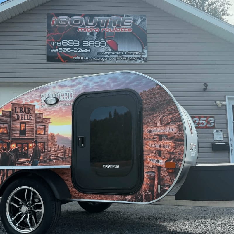 A teardrop trailer with a Western-themed wrap is parked in front of a building with a business sign.