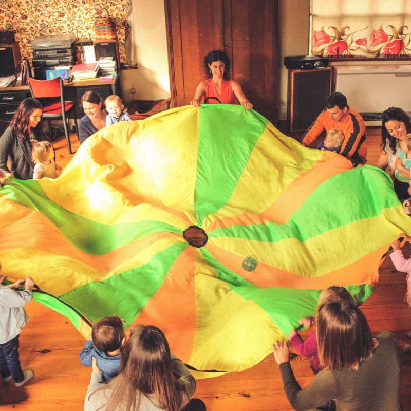 Children and adults are gathered around a large, colorful parachute in a room with wooden floors and a window in the background.