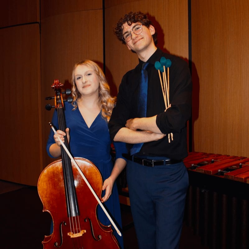A woman in a blue dress stands with a cello in the foreground, while a man in a black shirt and blue tie holds mallets in front of a marimba in the background.