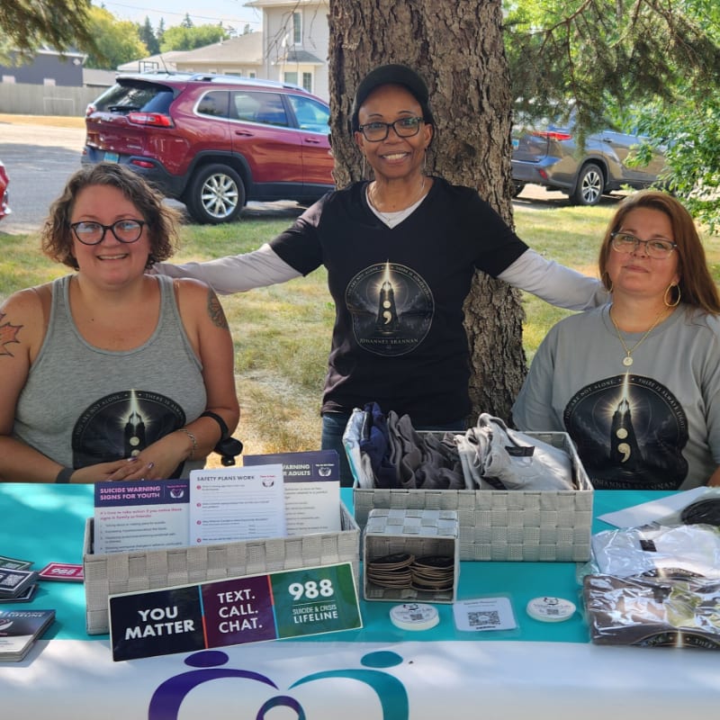Three women stand behind a table displaying mental health resources, with cars and trees visible in the background.