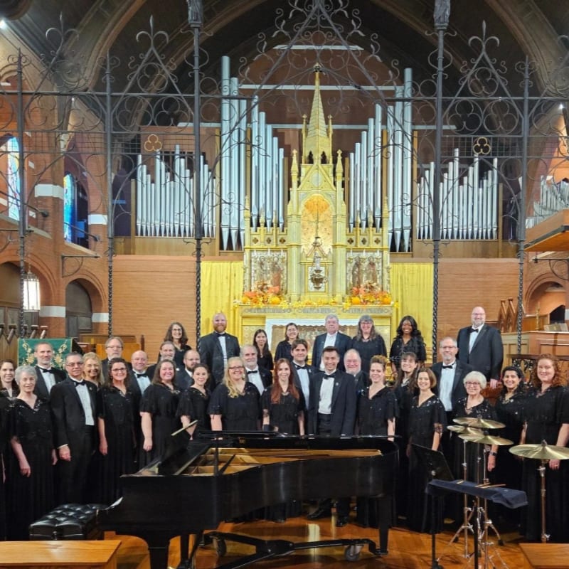 A choir stands in front of a grand pipe organ in a church, with a piano in the foreground.