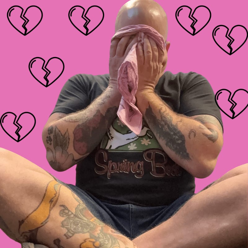 A bald man with tattoos on his arms and legs is sitting cross-legged and wiping his face with a pink cloth, surrounded by a pink background with broken heart graphics.